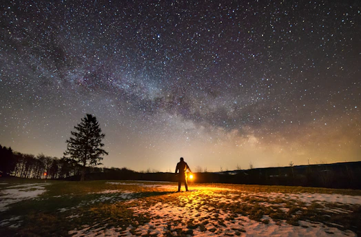 A solitary figure cloaked in gray, holding a glowing lantern, standing atop a snowy mountain under a starry night.