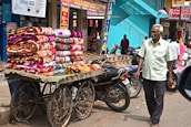 A street vendor pushes a cart loaded with colorful blankets, displaying intricate patterns. The setting is a bustling market street with motorcycles parked nearby, and pedestrians, including an elderly man in a light green shirt, walking alongside. The building behind has vibrant turquoise steps, with signage advertising festival offers.