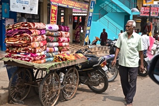 A street vendor pushes a cart loaded with colorful blankets, displaying intricate patterns. The setting is a bustling market street with motorcycles parked nearby, and pedestrians, including an elderly man in a light green shirt, walking alongside. The building behind has vibrant turquoise steps, with signage advertising festival offers.