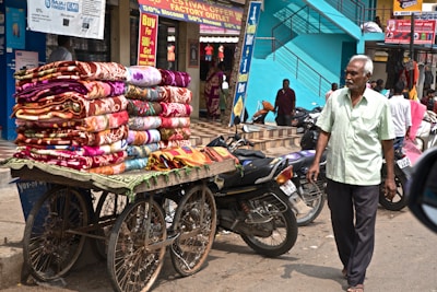 A street vendor pushes a cart loaded with colorful blankets, displaying intricate patterns. The setting is a bustling market street with motorcycles parked nearby, and pedestrians, including an elderly man in a light green shirt, walking alongside. The building behind has vibrant turquoise steps, with signage advertising festival offers.