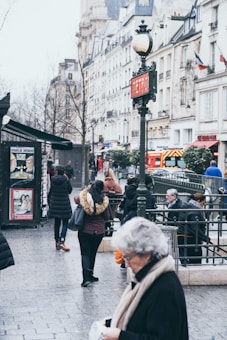 A bustling urban street scene depicts people walking near a metro entrance. The surroundings include a mix of modern and historical architecture. A sign labeled 'Metro' stands prominently, and there are shops and advertisements nearby. The atmosphere suggests a typical day in a European city with people going about their daily activities.