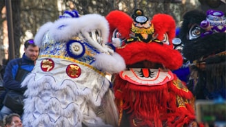 Two colorful lion costumes are seen, one white with blue and gold accents and the other red with intricate designs, worn by performers likely participating in a festive parade. They are surrounded by people observing the event.
