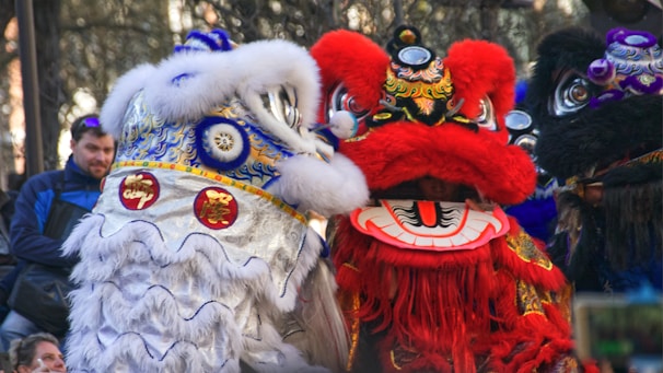Two colorful lion costumes are seen, one white with blue and gold accents and the other red with intricate designs, worn by performers likely participating in a festive parade. They are surrounded by people observing the event.