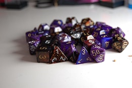 A pile of multi-sided dice, typically used in tabletop role-playing games, arranged on a smooth white surface. The dice are primarily in shades of purple, black, and dark brown, with numbers in contrasting colors, including gold and white, clearly visible on their faces.