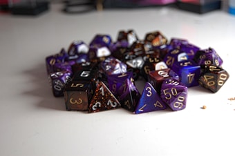 A pile of multi-sided dice, typically used in tabletop role-playing games, arranged on a smooth white surface. The dice are primarily in shades of purple, black, and dark brown, with numbers in contrasting colors, including gold and white, clearly visible on their faces.
