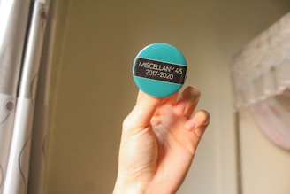 A hand holding a round badge with a teal background and a black stripe in the center. On the stripe, the text 'MISCELLANY 4.5 2017-2020' is printed in white. The background consists of light-colored curtains with a minimal pattern and a partial view of a white bed with draped fabric.