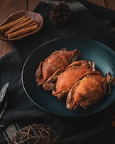 Three cooked crabs are arranged on a dark teal plate, set on a black cloth. To the side, a wooden bowl contains cinnamon sticks, and a pine cone is also visible. The setting has a rustic and cozy atmosphere.