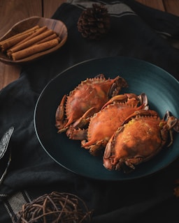 Three cooked crabs are arranged on a dark teal plate, set on a black cloth. To the side, a wooden bowl contains cinnamon sticks, and a pine cone is also visible. The setting has a rustic and cozy atmosphere.