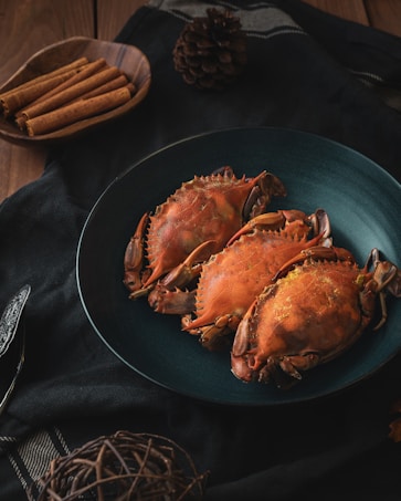 Three cooked crabs are arranged on a dark teal plate, set on a black cloth. To the side, a wooden bowl contains cinnamon sticks, and a pine cone is also visible. The setting has a rustic and cozy atmosphere.