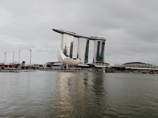 A prominent waterfront with a futuristic architectural complex comprising three interconnected high-rise buildings topped with a horizontal structure resembling a boat. In front of the buildings is a lotus-shaped structure adjacent to a body of water with reflections.