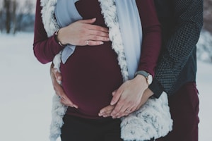 A couple is seen with their hands gently placed on a pregnant belly, highlighting a sense of anticipation and togetherness. The setting appears to be outdoors in a snowy landscape, with the woman wearing a burgundy top, a watch on her wrist, and a fur vest, while the man is wearing a dark dotted shirt. The image captures an intimate and serene moment.