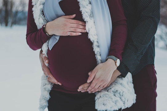 A couple is seen with their hands gently placed on a pregnant belly, highlighting a sense of anticipation and togetherness. The setting appears to be outdoors in a snowy landscape, with the woman wearing a burgundy top, a watch on her wrist, and a fur vest, while the man is wearing a dark dotted shirt. The image captures an intimate and serene moment.