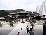 A traditional wooden temple complex surrounded by well-maintained gardens and pathways is situated against a backdrop of lush green hills. The foreground features several people walking, some taking photographs. In the distance, modern high-rise buildings can be seen towering over the landscape.
