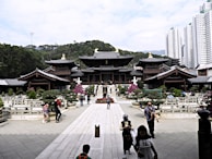 A traditional wooden temple complex surrounded by well-maintained gardens and pathways is situated against a backdrop of lush green hills. The foreground features several people walking, some taking photographs. In the distance, modern high-rise buildings can be seen towering over the landscape.