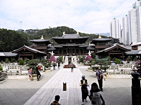 A traditional wooden temple complex surrounded by well-maintained gardens and pathways is situated against a backdrop of lush green hills. The foreground features several people walking, some taking photographs. In the distance, modern high-rise buildings can be seen towering over the landscape.