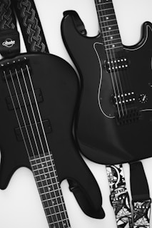 Two black guitars are visible, one bass guitar with four strings and one electric guitar with six strings. Both instruments rest against a light background, emphasizing their sleek design. The guitar straps have intricate patterns, adding a touch of personal style.