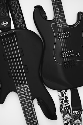 Two black guitars are visible, one bass guitar with four strings and one electric guitar with six strings. Both instruments rest against a light background, emphasizing their sleek design. The guitar straps have intricate patterns, adding a touch of personal style.