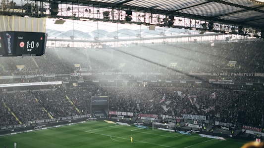 A large football stadium filled with spectators during a match. Sunlight filters through the transparent roof, illuminating parts of the stands and the field. A digital scoreboard displays a score of 1 to 0 at 46 minutes and 37 seconds. Banners and flags are visible among the dense crowd, adding to the vibrant atmosphere.