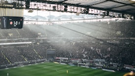 A large football stadium filled with spectators during a match. Sunlight filters through the transparent roof, illuminating parts of the stands and the field. A digital scoreboard displays a score of 1 to 0 at 46 minutes and 37 seconds. Banners and flags are visible among the dense crowd, adding to the vibrant atmosphere.