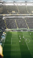 A crowded stadium with fans fills the stands surrounding a soccer field. On the field, players are actively engaged, with some in white jerseys and others in dark jerseys, moving toward a goal where a goalkeeper is visible.