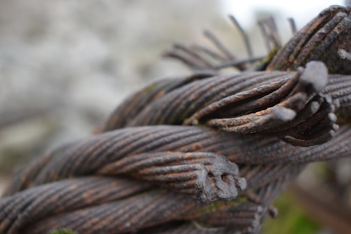 A close-up of twisted and corroded metal cables intertwined with each other. The metallic surface shows signs of rust and decay, suggesting it has been exposed to the elements for a long time. The background is blurred, drawing attention to the detailed texture of the metal strands.