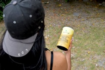 A person wearing a dark cap and tank top is holding a yellow container labeled 'Organic Life Rice Protein' in an outdoor setting. The ground is covered with grass and scattered leaves, creating a natural environment.