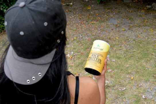 A person wearing a dark cap and tank top is holding a yellow container labeled 'Organic Life Rice Protein' in an outdoor setting. The ground is covered with grass and scattered leaves, creating a natural environment.