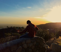 A person wearing a red plaid shirt sits on a rock wall playing a guitar, overlooking a scenic view at sunset. The sun casts warm hues across the landscape, highlighting a sign nearby that reads 'No Smoking or Fires'. The sky transitions from blue to orange as it approaches the horizon.