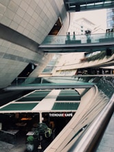 A modern architectural setting with clean lines and curved structures, featuring a multi-level space with signs for 'Kesfet Intema Yasam' and 'Kanyon'. The floor level has a café named 'The House Café' with green awnings. Vegetation adds a touch of nature to the urban environment.