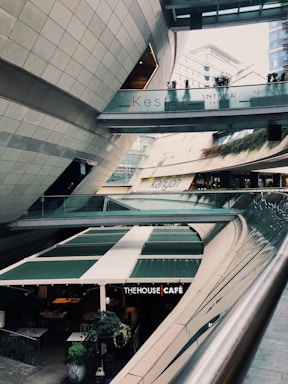 A modern architectural setting with clean lines and curved structures, featuring a multi-level space with signs for 'Kesfet Intema Yasam' and 'Kanyon'. The floor level has a café named 'The House Café' with green awnings. Vegetation adds a touch of nature to the urban environment.