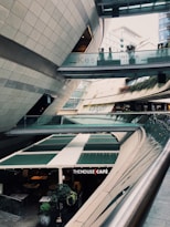 A modern architectural setting with clean lines and curved structures, featuring a multi-level space with signs for 'Kesfet Intema Yasam' and 'Kanyon'. The floor level has a café named 'The House Café' with green awnings. Vegetation adds a touch of nature to the urban environment.