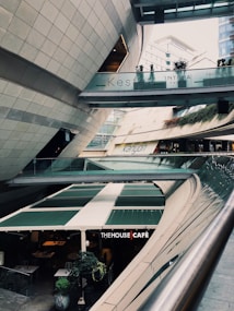 A modern architectural setting with clean lines and curved structures, featuring a multi-level space with signs for 'Kesfet Intema Yasam' and 'Kanyon'. The floor level has a caf&eacute; named 'The House Caf&eacute;' with green awnings. Vegetation adds a touch of nature to the urban environment.