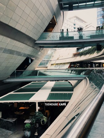 A modern architectural setting with clean lines and curved structures, featuring a multi-level space with signs for 'Kesfet Intema Yasam' and 'Kanyon'. The floor level has a caf&eacute; named 'The House Caf&eacute;' with green awnings. Vegetation adds a touch of nature to the urban environment.