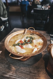 A brass pan containing a steaming dish with white fluffy elements and garnished with sliced green and red peppers sits on a wooden table. The background shows a dimly lit restaurant interior with chairs and people in casual attire.