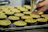 A hand is seen filling uncooked tart shells with a creamy yellow mixture using a piping bag. The tarts are arranged on a metal baking tray.
