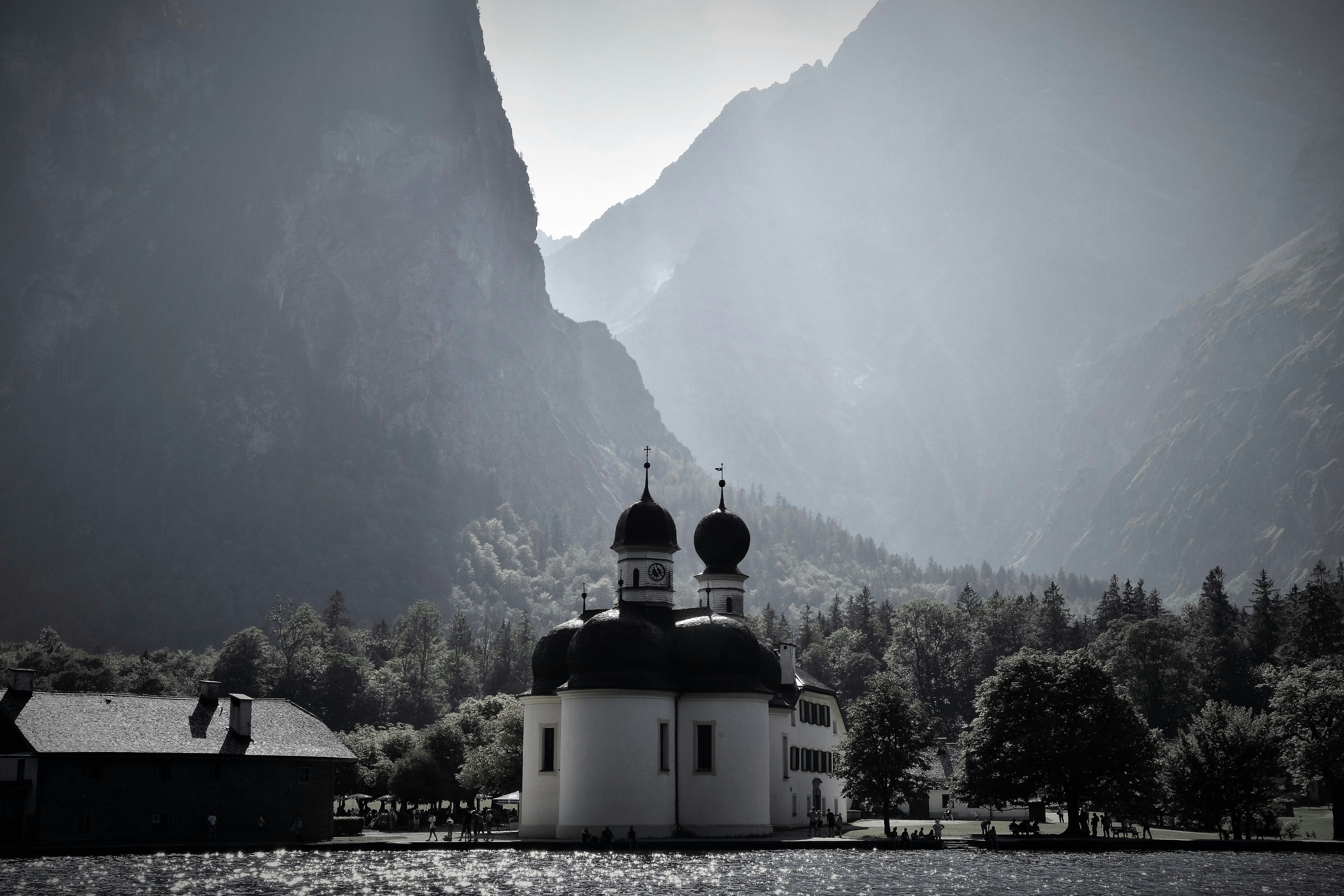 grayscale photo of chapel near body of water surrounded with mountains