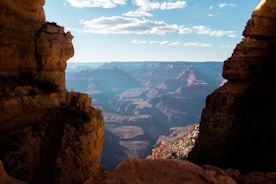 The image displays a vast canyon landscape under a clear blue sky, framed by rugged, rocky cliffs on both sides. The distant canyon walls are illuminated by sunlight, highlighting their layered formations and reddish hues.