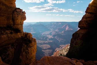 The image displays a vast canyon landscape under a clear blue sky, framed by rugged, rocky cliffs on both sides. The distant canyon walls are illuminated by sunlight, highlighting their layered formations and reddish hues.