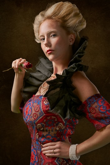 A person wearing an elegant, vintage-style dress decorated with intricate lace patterns in red and blue. The dress features puffed shoulders and is accessorized with pearl jewelry including a necklace and bracelet. They hold a red rose close to their face. The hairstyle is elaborate and reminiscent of historical fashion, with hair styled high above the head.