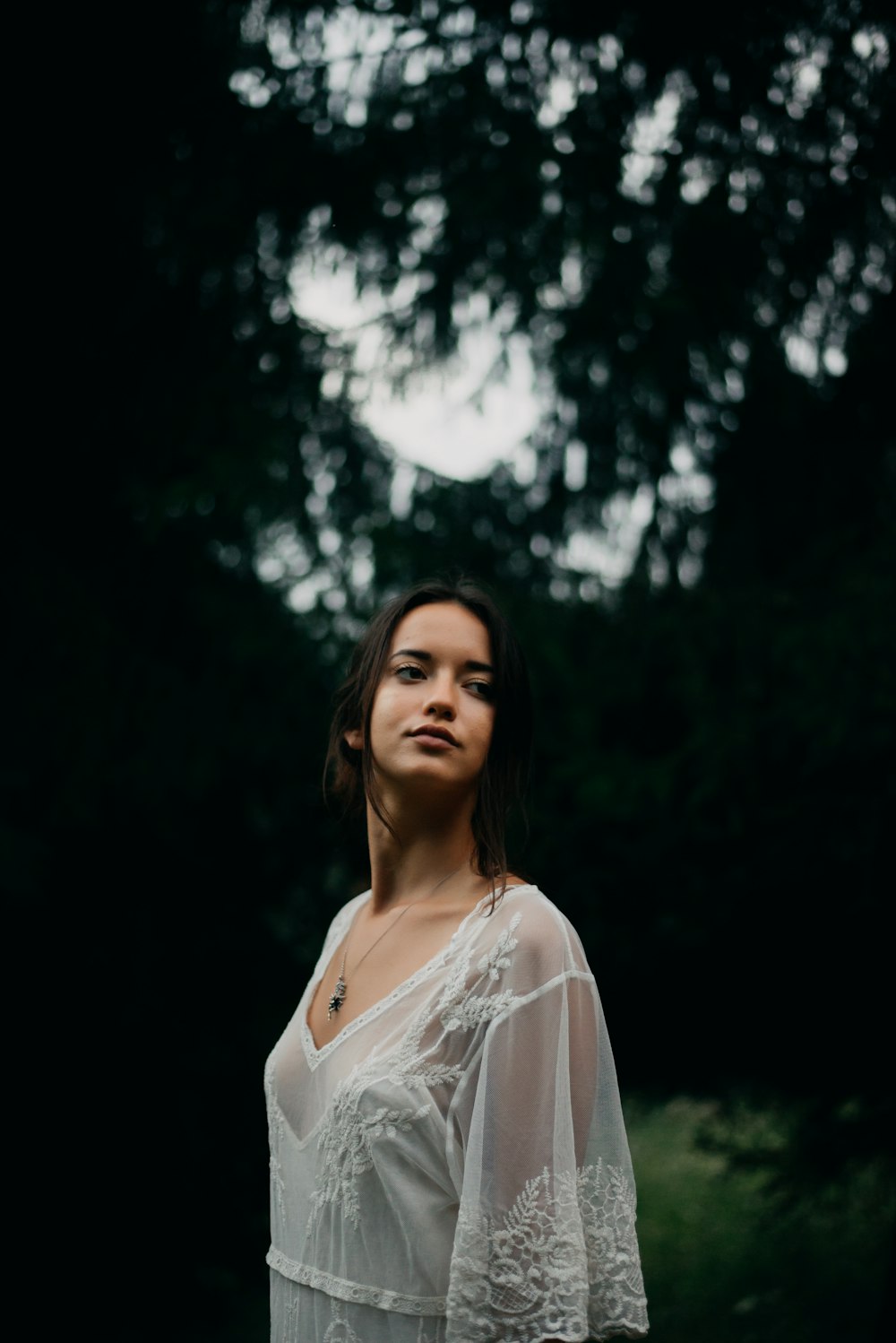 selective focus photography of woman standing near trees wearing white sheer top