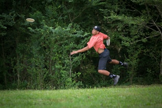 A person in a bright orange shirt and dark shorts is jumping in mid-air while throwing a frisbee in a grassy outdoor area surrounded by dense green foliage.