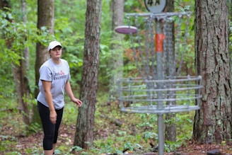 A person is playing disc golf in a forested area, focusing on throwing a disc towards a metal basket. The scene is set amongst tall trees with lush green foliage.