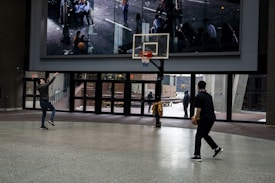 Two individuals are playing basketball in an indoor setting with a large digital screen in the background. The floor is tiled, and there is a wall of windows that look out to an outdoor area with a few people visible. One player is shooting towards the hoop while the other is holding a basketball, preparing to play.