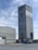 A tall, rectangular industrial building with cables extending from the top. The structure is made of concrete and features a logo with the words 'Fresnillo Saucito'. Adjacent to it is a lower building with a corrugated metal roof and a stone facade. In the foreground, there is construction equipment, including a blue truck and a loader under a clear sky with some clouds.