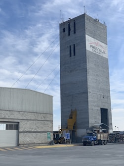 A tall, rectangular industrial building with cables extending from the top. The structure is made of concrete and features a logo with the words 'Fresnillo Saucito'. Adjacent to it is a lower building with a corrugated metal roof and a stone facade. In the foreground, there is construction equipment, including a blue truck and a loader under a clear sky with some clouds.