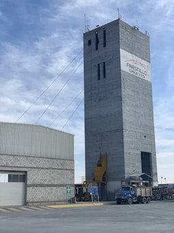 A tall, rectangular industrial building with cables extending from the top. The structure is made of concrete and features a logo with the words 'Fresnillo Saucito'. Adjacent to it is a lower building with a corrugated metal roof and a stone facade. In the foreground, there is construction equipment, including a blue truck and a loader under a clear sky with some clouds.