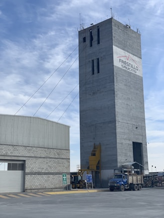 A tall, rectangular industrial building with cables extending from the top. The structure is made of concrete and features a logo with the words 'Fresnillo Saucito'. Adjacent to it is a lower building with a corrugated metal roof and a stone facade. In the foreground, there is construction equipment, including a blue truck and a loader under a clear sky with some clouds.
