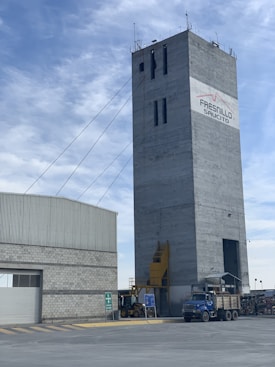 A tall, rectangular industrial building with cables extending from the top. The structure is made of concrete and features a logo with the words 'Fresnillo Saucito'. Adjacent to it is a lower building with a corrugated metal roof and a stone facade. In the foreground, there is construction equipment, including a blue truck and a loader under a clear sky with some clouds.