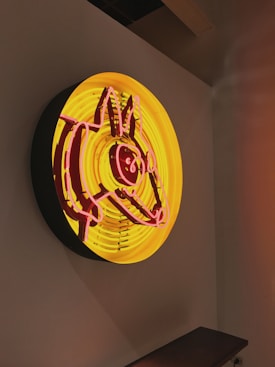 A brightly lit neon sign features an abstract drawing of a cat with prominent ears and facial features in pink lines over a yellow circular background. The sign is mounted on a plain wall, creating a striking contrast between the vibrant colors and the neutral surroundings.