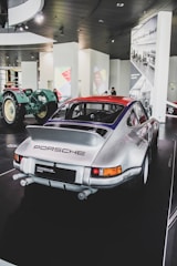 A classic silver Porsche vehicle is displayed in what appears to be a modern car museum. The car is parked on a black, glossy floor, and its rear end is prominently visible, showcasing the Porsche logo and detailing. In the background, a vintage green tractor adds contrast and historical context. The walls feature bright graphic panels and black-and-white photographs, adding visual diversity.